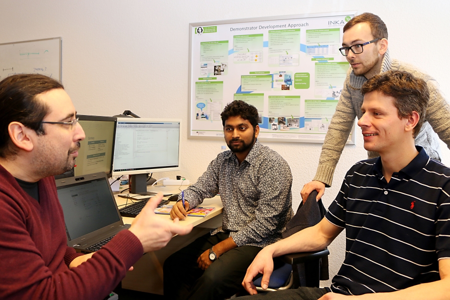 Vier Männer sind in einem Büro vor einem Computer und unterhalten sich.